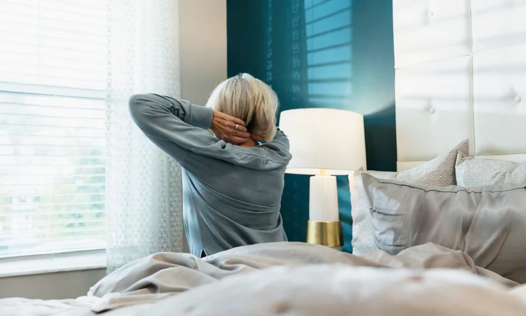 Woman stretching her neck beside a bed for cervical pain relief.