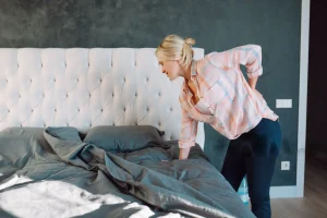 Woman with lower back pain making a bed, highlighting the choice between an adjustable bed and orthopedic mattress.